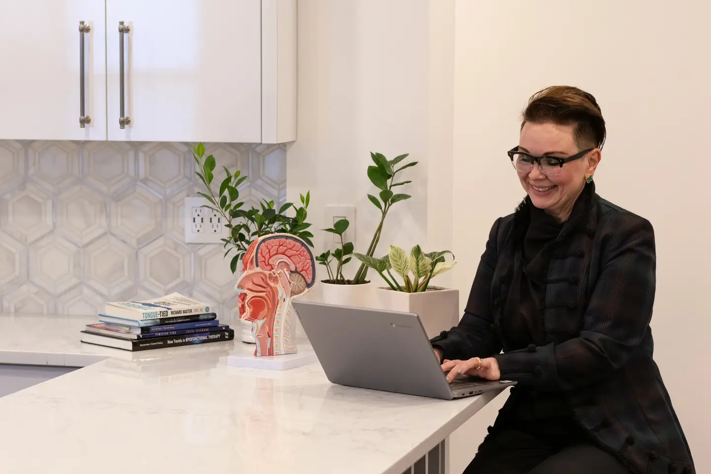 Julia Yarovoy leading an online myofunctional therapy session on a laptop, with an anatomical airway model and myofunctional therapy books beside her